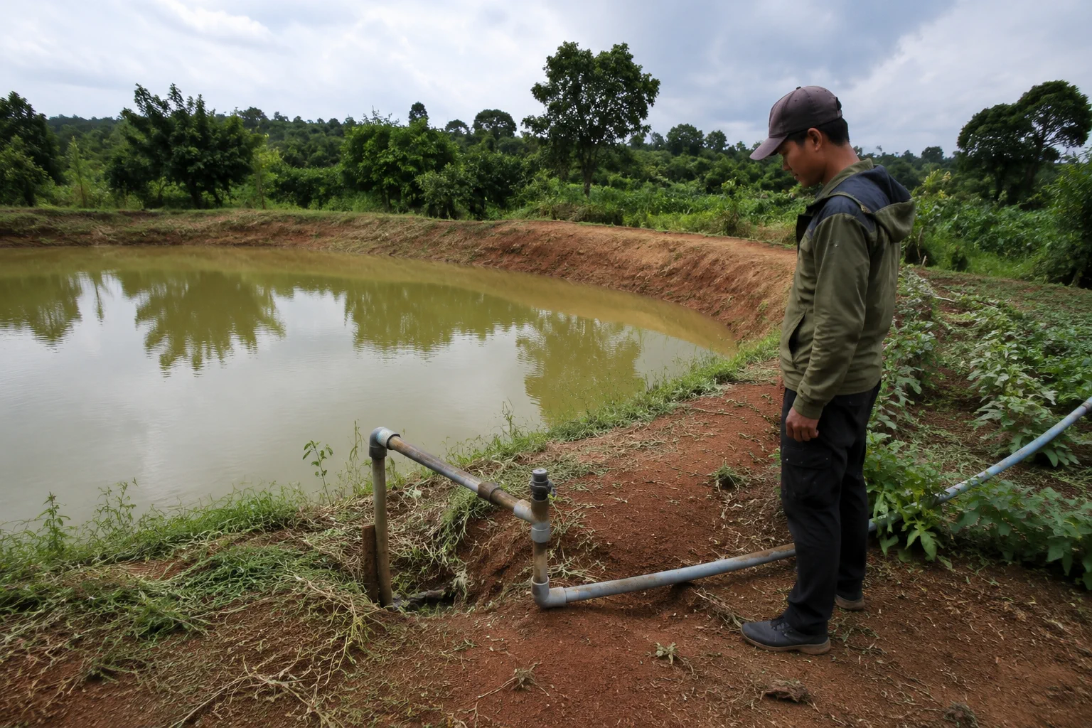 Toàn cảnh một khu đất nông nghiệp tại Đắk Nông với người khảo sát đang kiểm tra thực tế nguồn nước tại một ao chứa nhỏ cạnh khu canh tác, quan sát đường ống tưới và khu vực xung qu